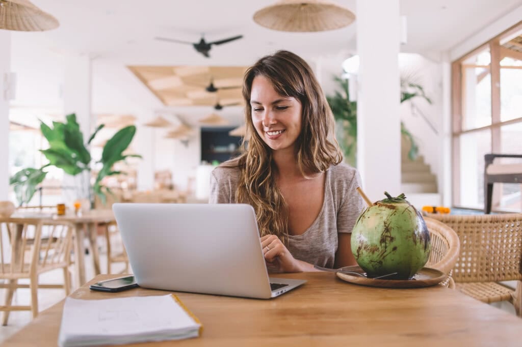 Cheerful woman in casual wear creating excited article with information about tropical fruits sitting at cafeteria in Bali and connecting to public internet, positive hipster girl enjoying chatting